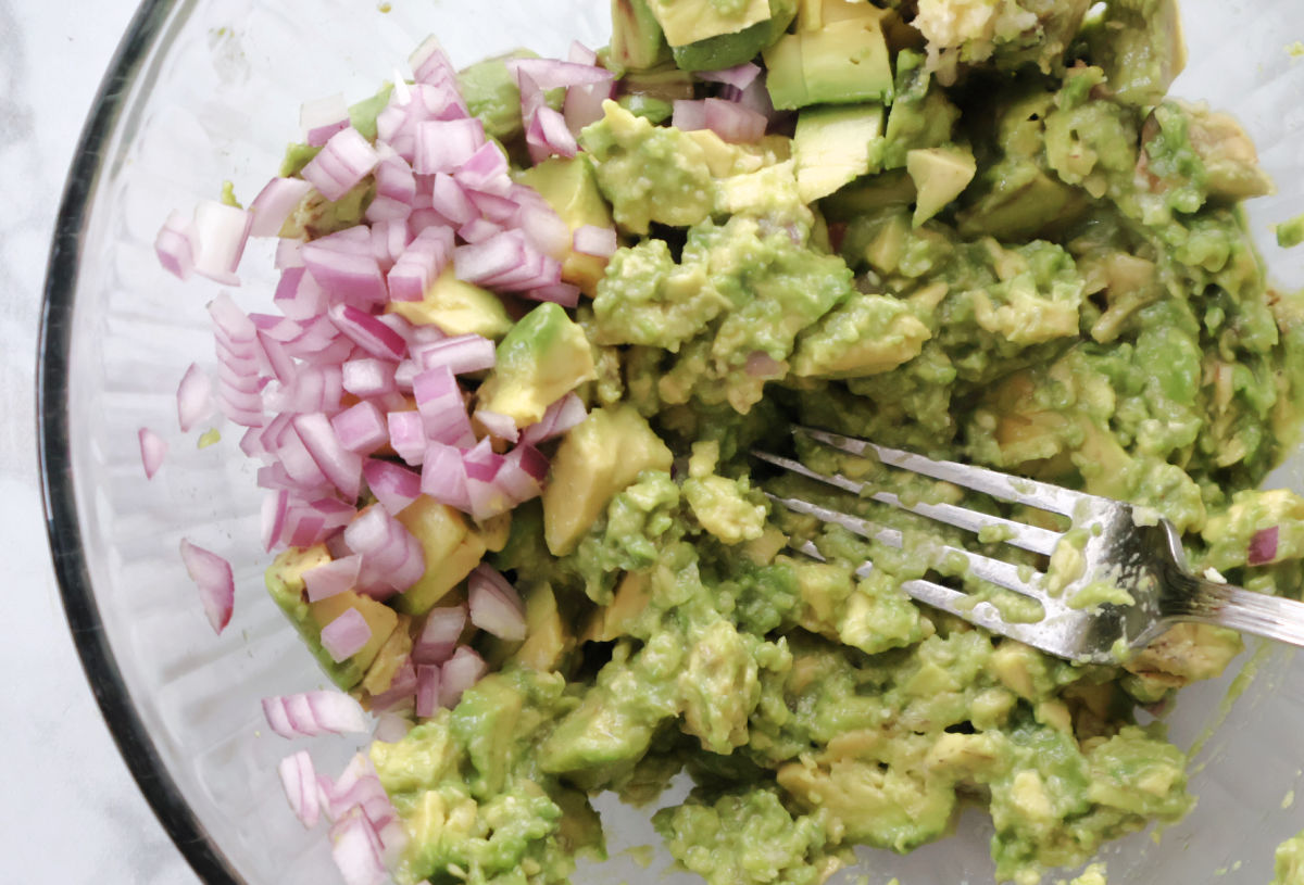 avocado being mashed and red onions added to recipe for fresh guacamole,