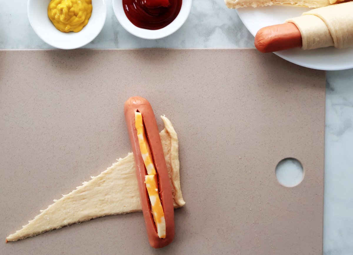 A hot dog wiener with a slit filled with cheese.  It is lying on the wide side of a crescent roll triangle.