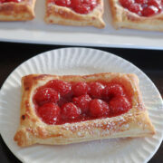 cherry danish with a dusting of powdered sugar. More cherry danishes behind the single danish on a tray.