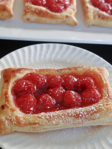 cherry danish with a dusting of powdered sugar. More cherry danishes behind the single danish on a tray.