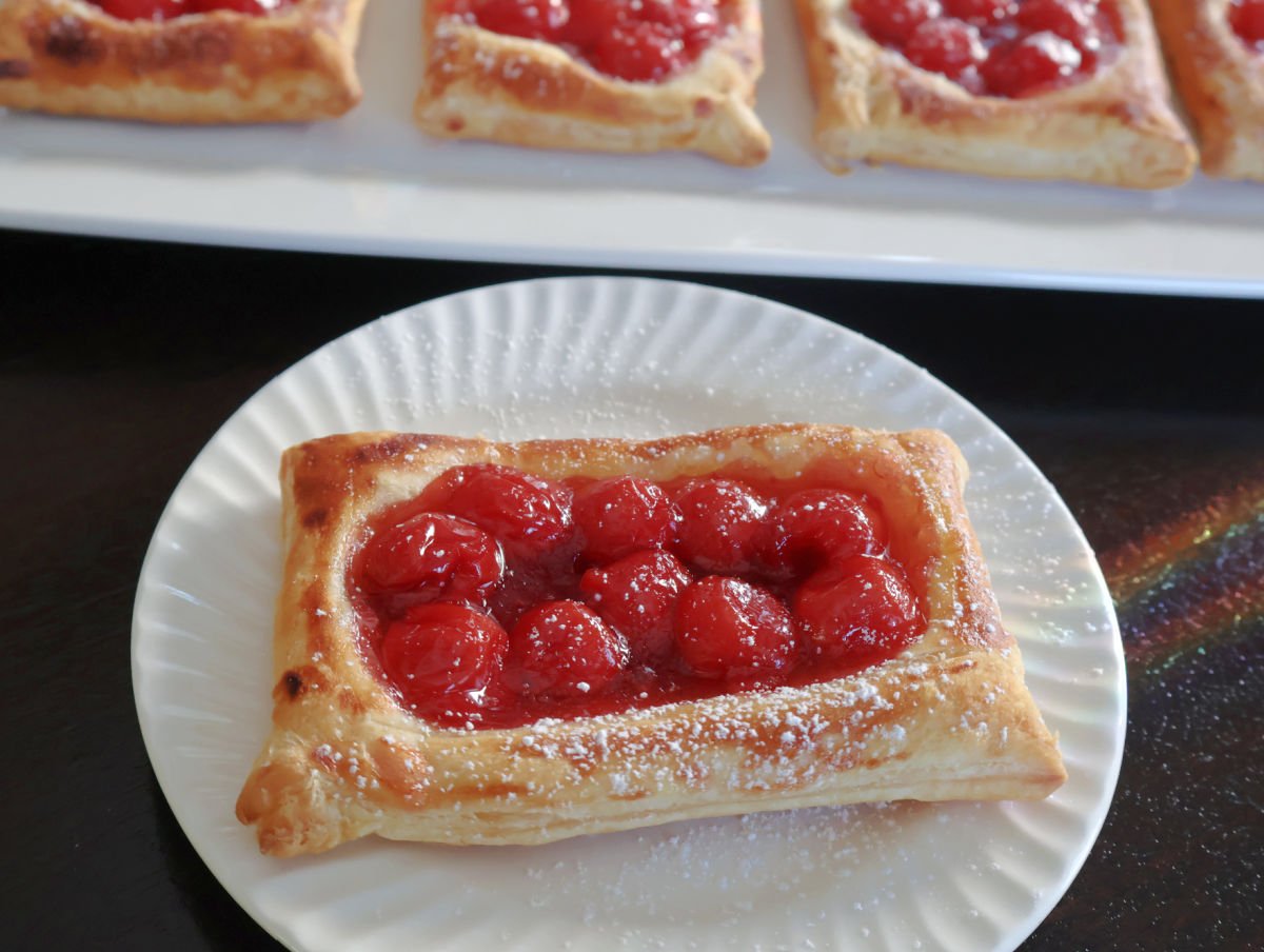 cherry danish with a dusting of powdered sugar. More cherry danishes behind the single danish on a tray.