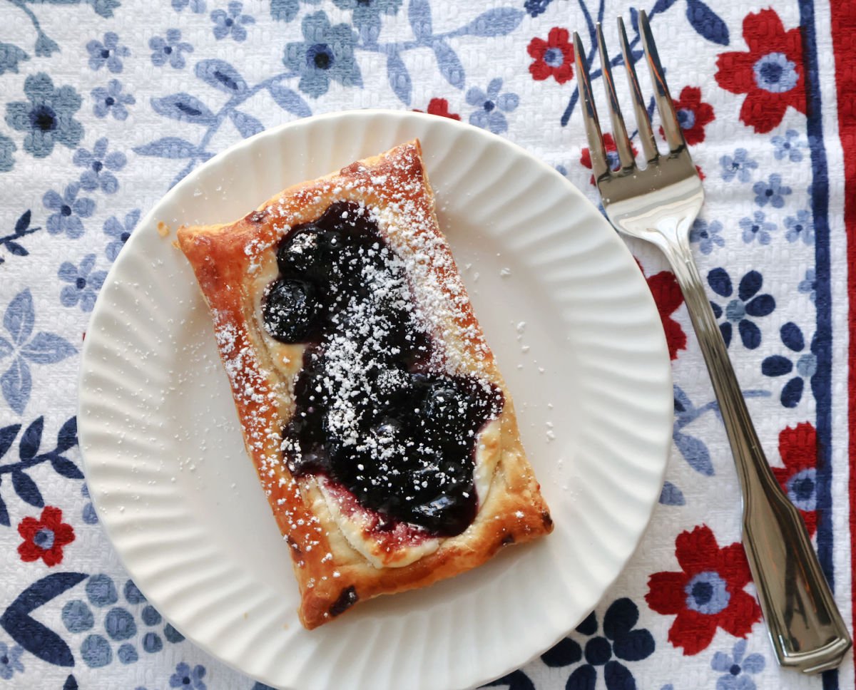 blueberry cheese danish on a white plate, placed on a tablecloth with blue and red small flowers.