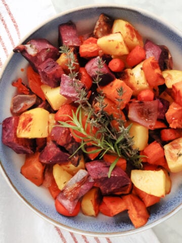 Roasted sweet potatoes in a serving bowl. Includes roasted potatoes, sweet potatoes, purple sweet potatoes, carrots and red onions. Seasoned then garnished with a sprig of rosemary and some thyme.