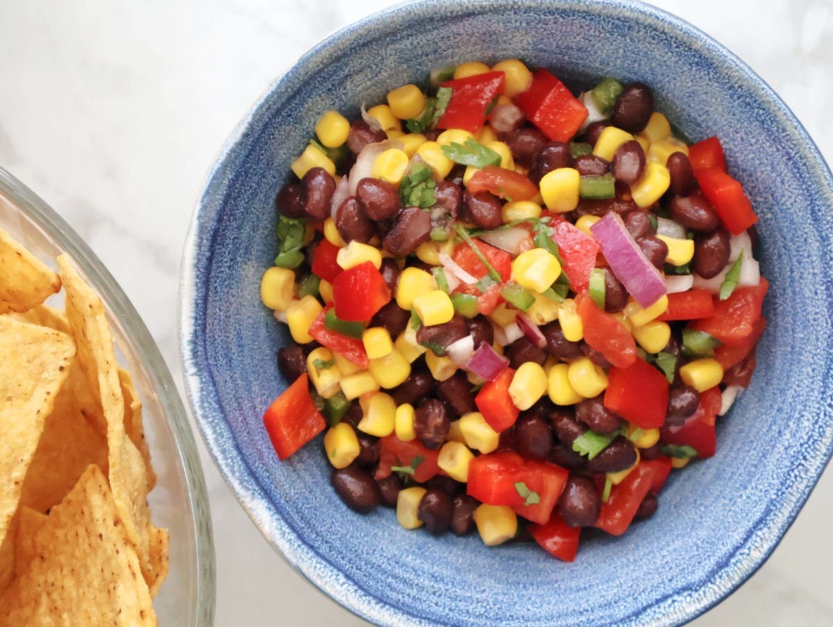 Black beans, corn, diced tomatoes, and red onion in a blue serving bowl after tossing with dressing. Corn tortilla chips are in a bowl to the side.