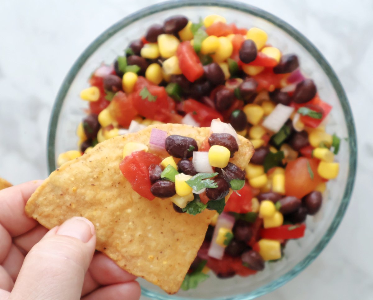 Scoop of black bean and corn salsa on a chip held above a white bowl, ready to eat