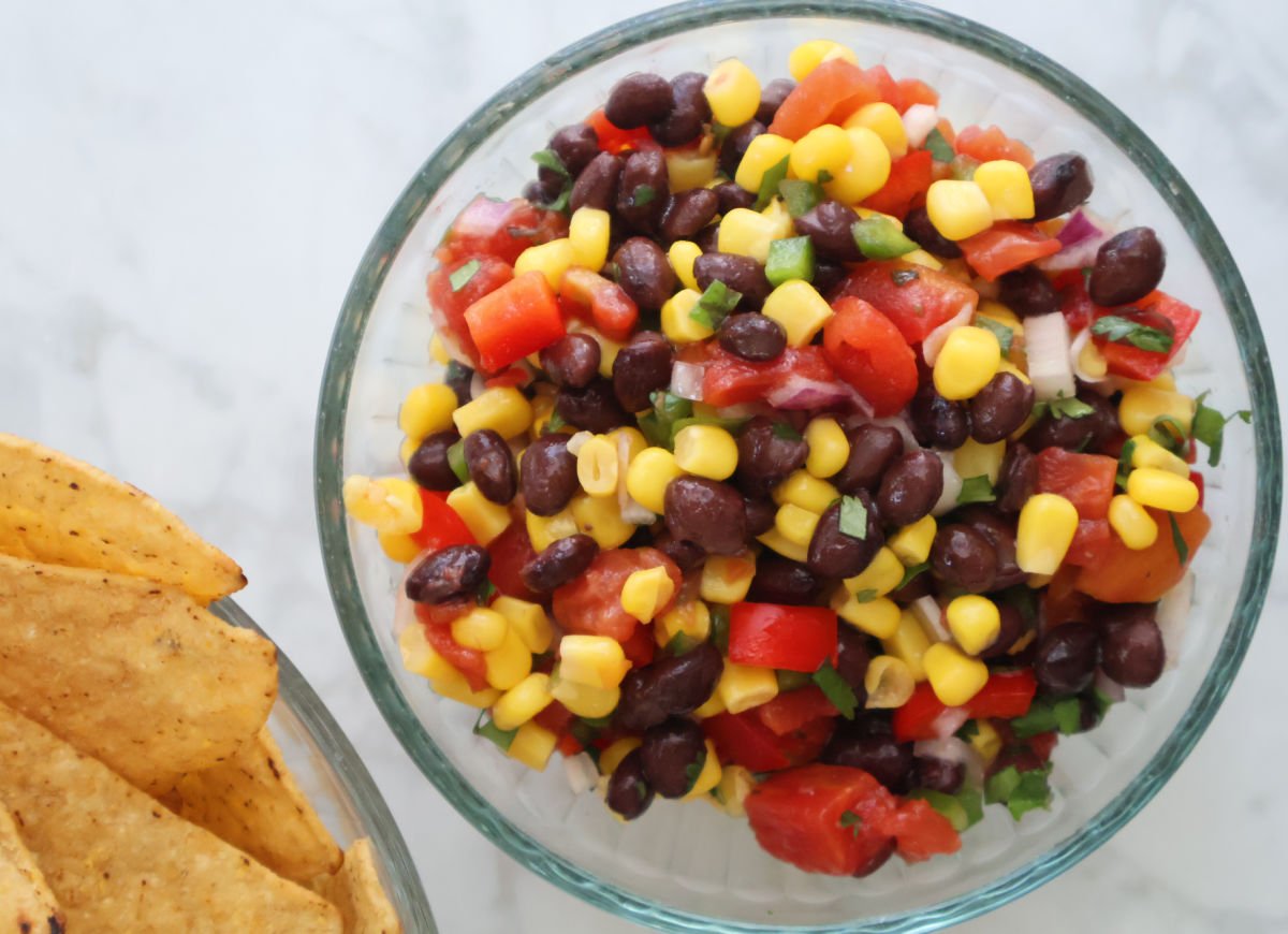 Black beans, corn, diced tomatoes, and red onion in a glass mixing bowl after tossing with dressing. Corn tortilla chips are in a bowl to the side.