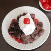 A serving of chocolate cherry dump cake on a white plate, topped with whipped cream and a maraschino cherry.