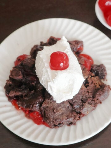 A serving of chocolate cherry dump cake on a white plate, topped with whipped cream and a maraschino cherry.