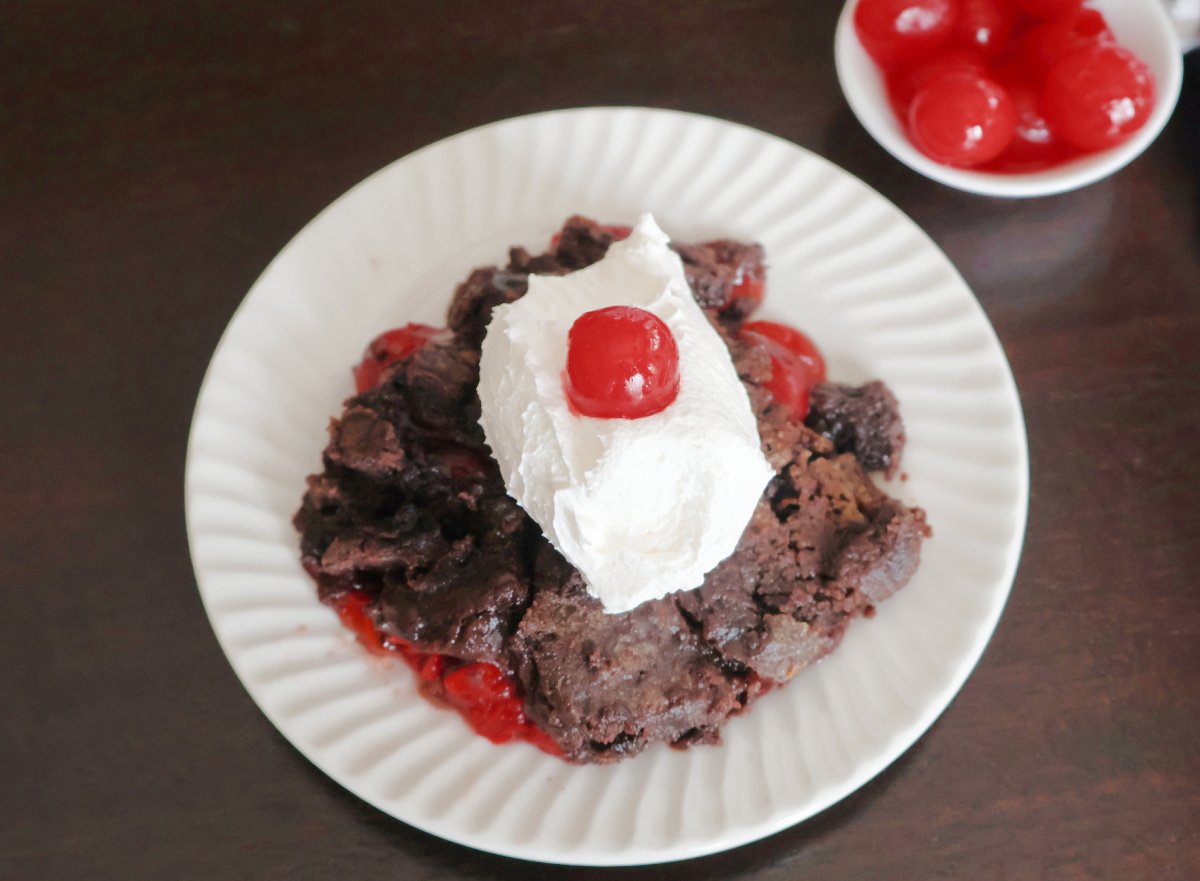 A serving of chocolate cherry dump cake on a white plate, topped with whipped cream and a maraschino cherry.