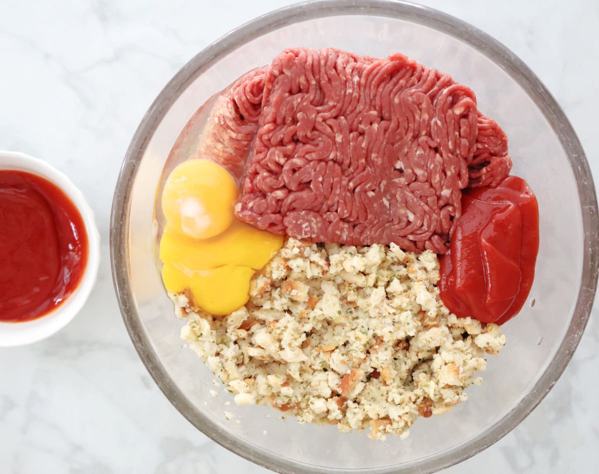 Ingredients photo showing ground beef in a bowl with eggs, ketchup bottle, and box of stuffing mix nearby - everything needed for this easy meatloaf recipe