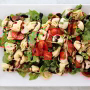 A fresh and colorful tortellini Caprese salad with cherry tomatoes, mozzarella balls, baby spinach, and basil, drizzled with balsamic glaze. Served cold as a summer pasta salad.