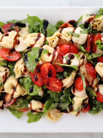 A fresh and colorful tortellini Caprese salad with cherry tomatoes, mozzarella balls, baby spinach, and basil, drizzled with balsamic glaze. Served cold as a summer pasta salad.
