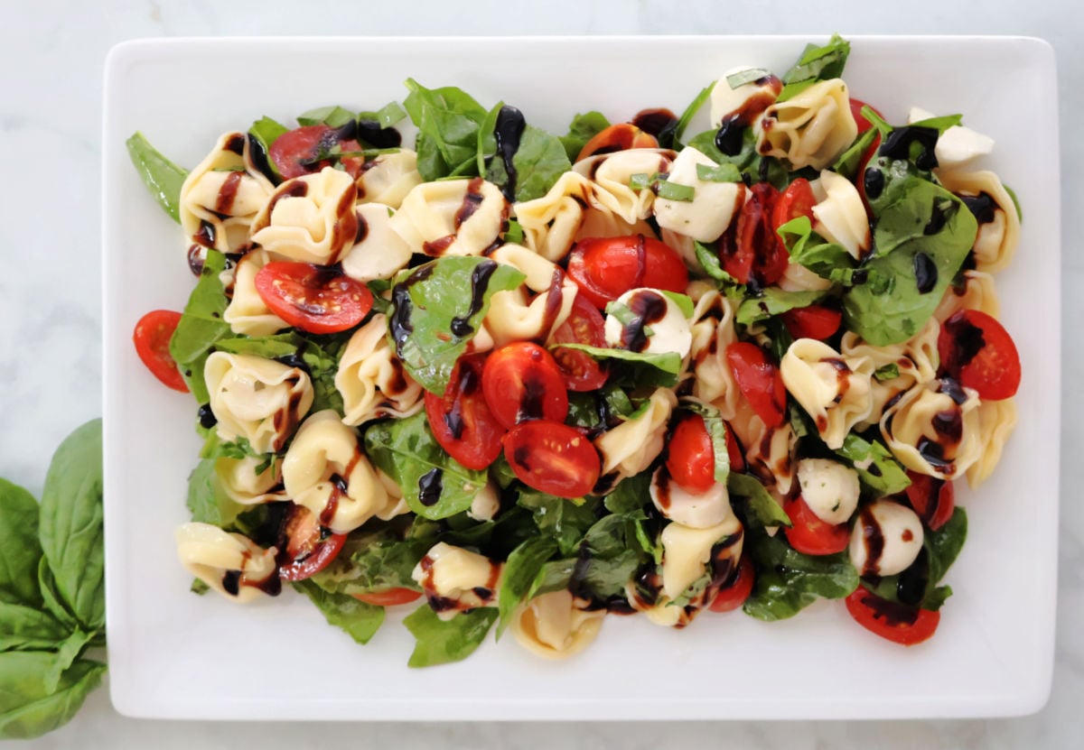 A fresh and colorful tortellini Caprese salad with cherry tomatoes, mozzarella balls, baby spinach, and basil, drizzled with balsamic glaze. Served cold as a summer pasta salad.