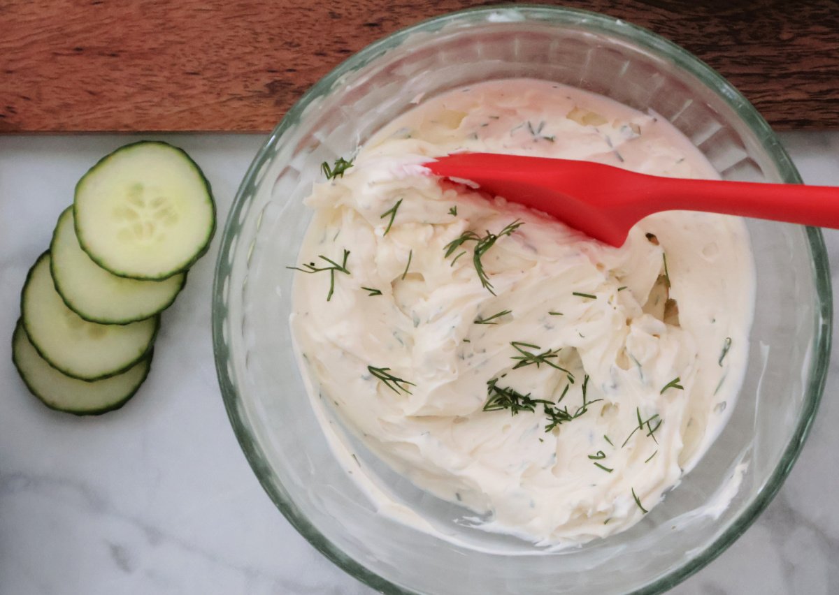 Stirring together cream cheese, ranch dressing, mayo and fresh dill using a red spoon.