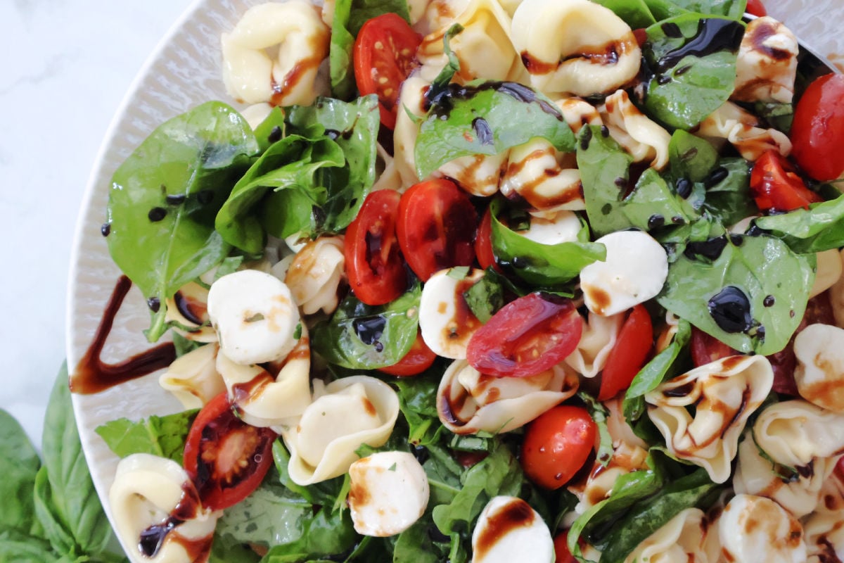 A fresh and colorful tortellini Caprese salad with cherry tomatoes, mozzarella balls, baby spinach, and basil, drizzled with balsamic glaze. Served cold as a summer pasta salad.