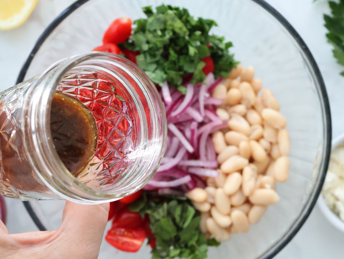 dressing in a mason jar being poured over cannellini beans and tomato salad ingredients