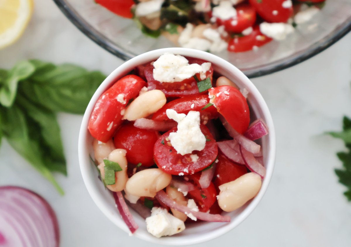 Overhead view of white bean and tomato salad with feta in a white bowl, garnished with fresh basil and parsley.