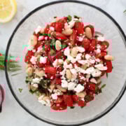 Overhead view of white bean and tomato salad with feta in a glass bowl, garnished with fresh basil and parsley.