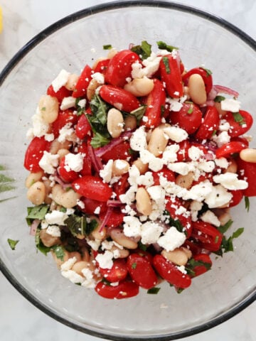 Overhead view of white bean and tomato salad with feta in a glass bowl, garnished with fresh basil and parsley.