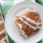 zucchini cake serving with icing glaze. On a white plate with a baking dish with zucchini cake in the background.