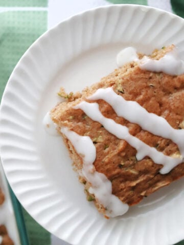 zucchini cake serving with icing glaze. On a white plate with a baking dish with zucchini cake in the background.