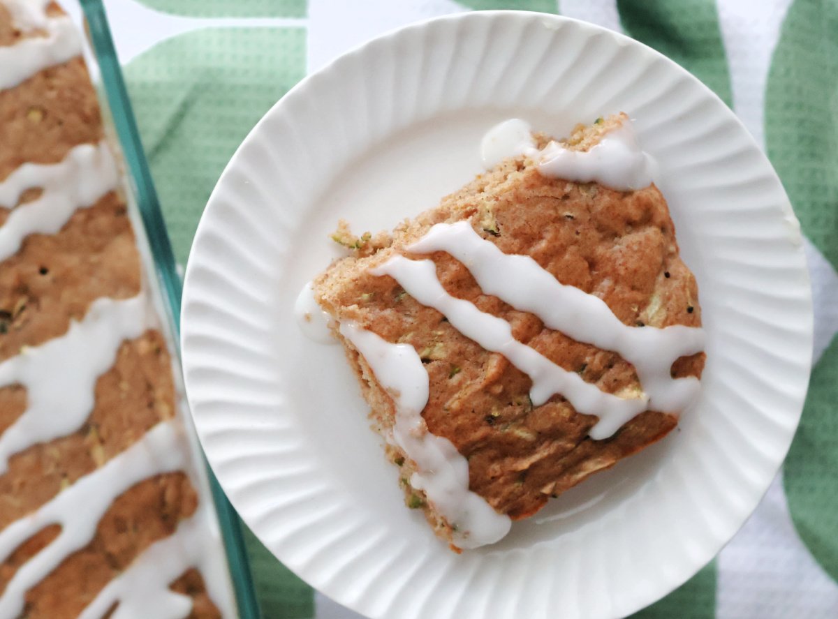 zucchini cake serving with icing glaze. On a white plate with a baking dish with zucchini cake in the background.