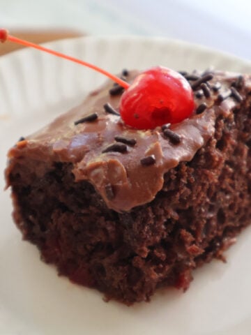 A piece of dr pepper cake with boiled icing garnished with a marachino cherry.