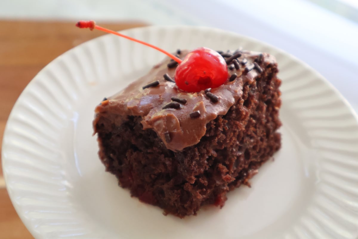A piece of dr pepper cake with boiled icing garnished with a marachino cherry.