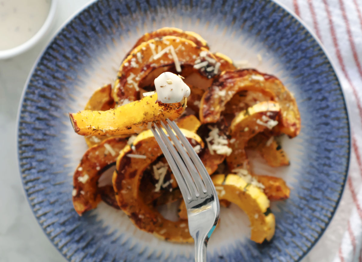 Delacata half moons, roasted and seasoned on a blue plate.  A fork has a delicata fry on it that has been dipped in ranch dressing.