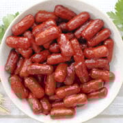 Appetizer sausages coated in grape jelly chili sauce served in a white bowl with toothpicks for easy snacking.