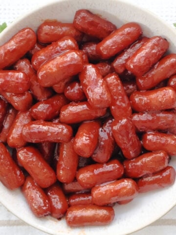 Appetizer sausages coated in grape jelly chili sauce served in a white bowl with toothpicks for easy snacking.