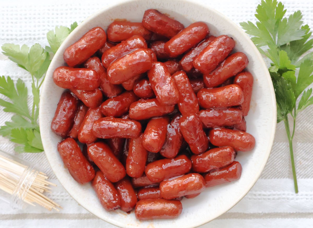 Appetizer sausages coated in grape jelly chili sauce served in a white bowl with toothpicks for easy snacking.