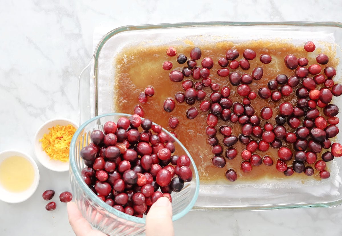 Fresh cranberries sprinkled with orange zest and juice over brown sugar and butter layer in baking dish.