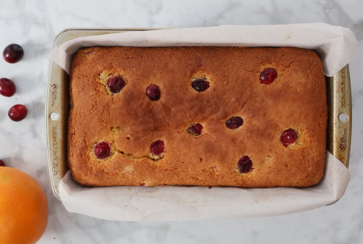 freshly baked cranberry orange bread in a loaf pan. Fresh cranberries and an orange sit to the left of the loaf pan as related ingrediets.