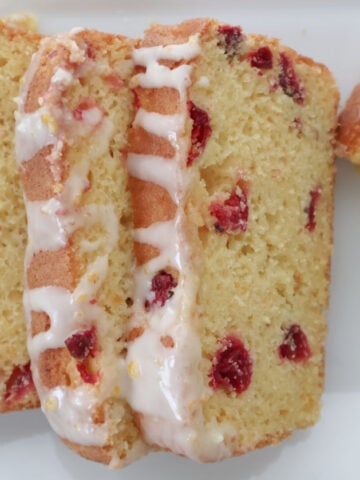 Slice of cranberry orange bread on a white plate showing tart cranberries and bright orange glaze drizzled on a few slices