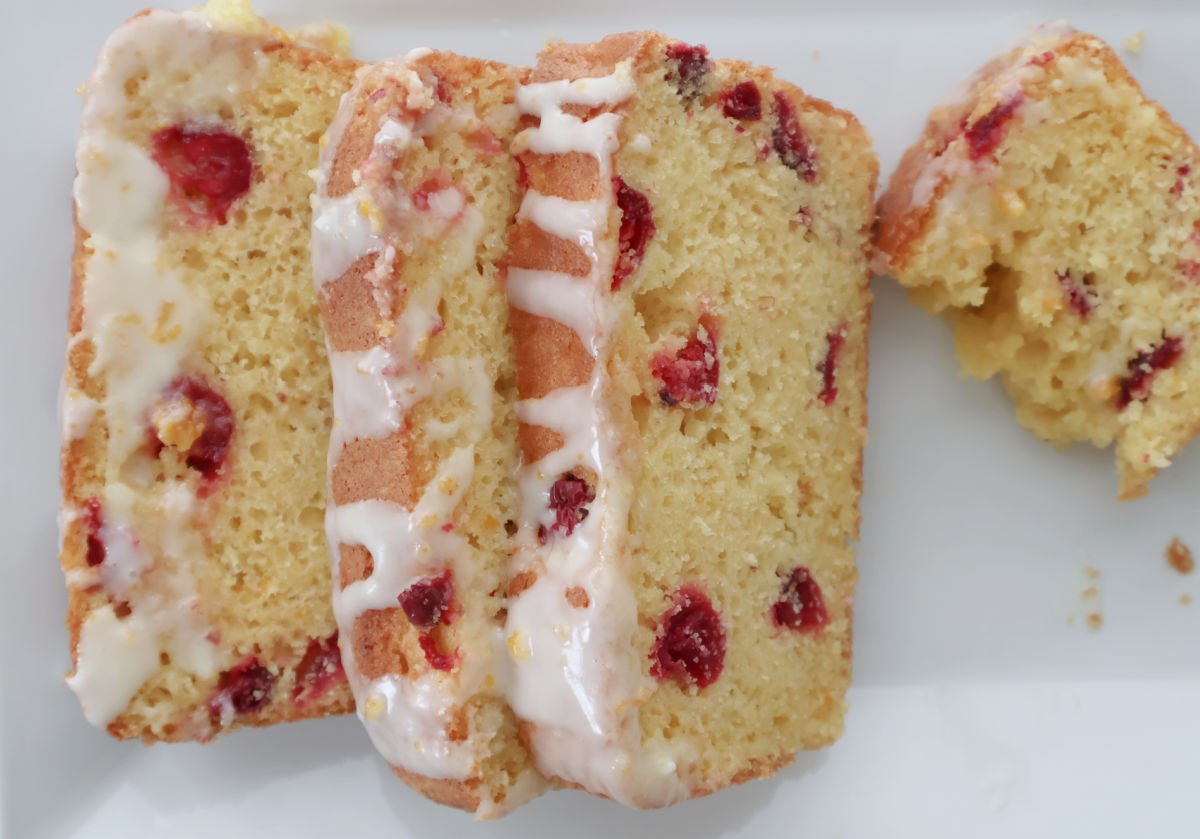 Slice of cranberry orange bread on a white plate showing tart cranberries and bright orange glaze drizzled on a few slices