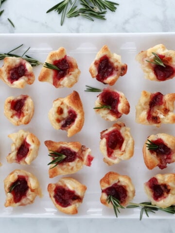 cranberry brie bites displayed on a white tray with rosemary garnish on the tray.